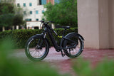 A 26" Step Over Electric Mountain Bike - Black from Evolvate Pro is parked outside on a paved surface near a building. The bike showcases thick, rugged tires, a sturdy frame, a swappable battery design, and a small yellow design element. The BAFANG rear hub motor ensures smooth rides. Greenery and other buildings are visible in the background.