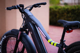 A close-up of a 26" Step Over Electric Mountain Bike - Black by Evolvate Pro showcases its thick tires and sleek, angular frame with an LCD touchscreen on the handlebars. The bike features a BAFANG rear hub motor and is set against a slightly blurred background of paved ground and greenery, giving the scene a clean and modern look.
