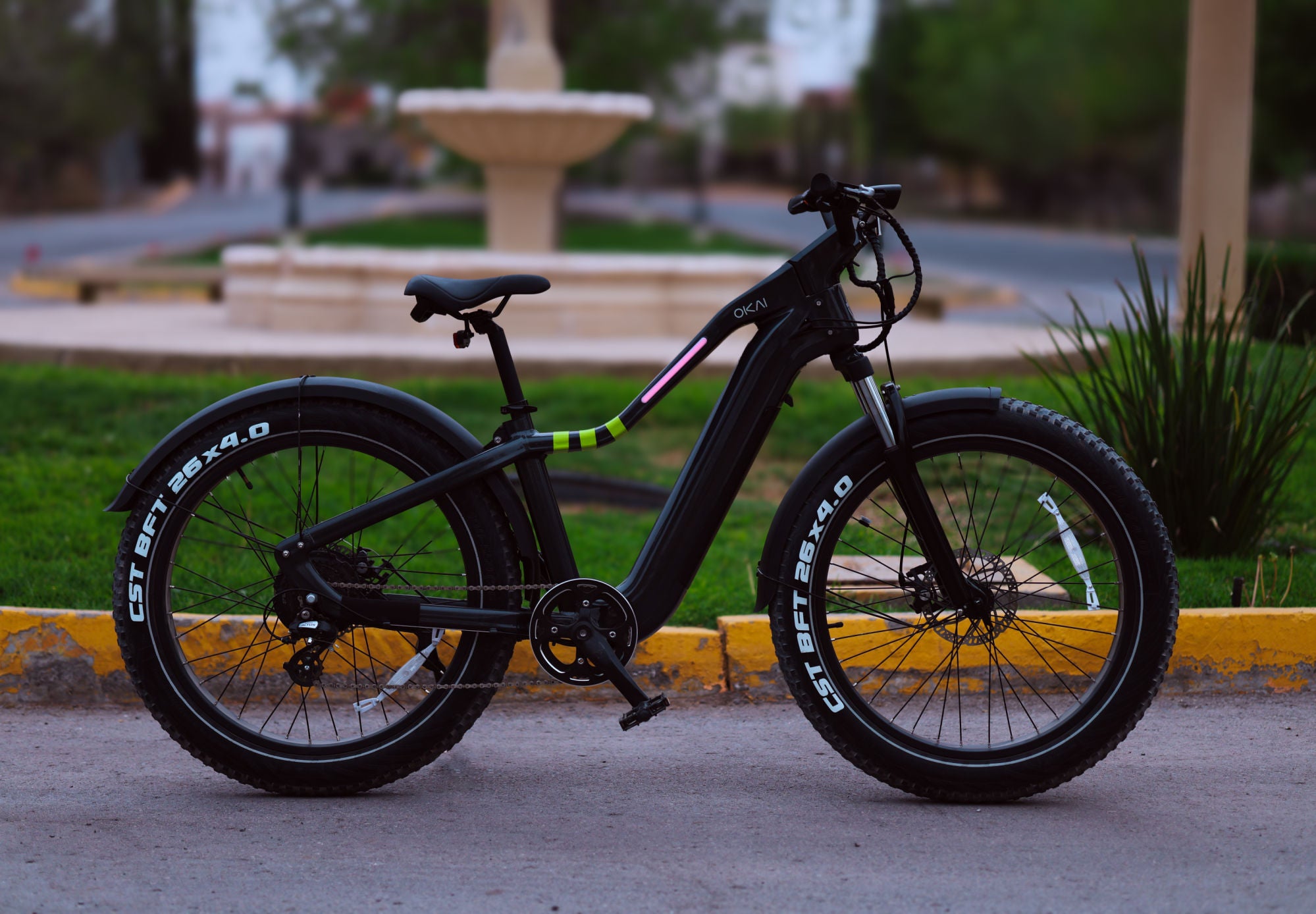 An Evolvate Pro 26" Step Over Electric Mountain Bike in black, featuring large tires, is parked on a paved path beside a grassy area and a fountain. The bike boasts a sturdy frame, customizable lighting bar, and visible battery pack. Equipped with a BAFANG rear hub motor, road markings are slightly visible in the background.