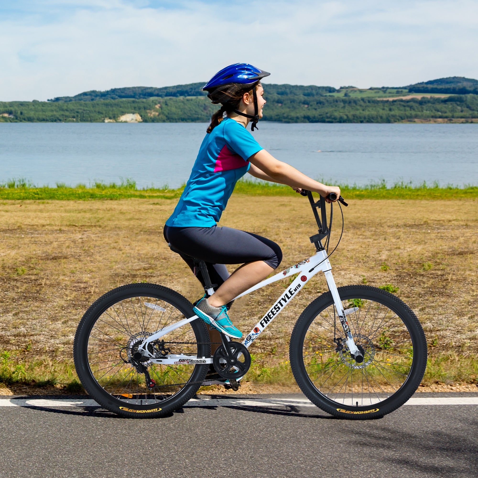 A person wearing a blue helmet, blue shirt, black shorts, and blue shoes is riding a purple Evolvate Pro Freestyle Kids Bike labeled "FREESTYLE" on a road. Equipped with Shimano 7-Speed gears and double disc brakes, the bike glides smoothly. A body of water and rolling hills are visible in the background, creating a peaceful scene.